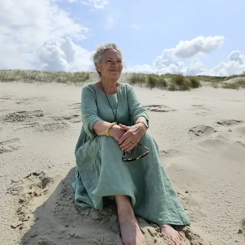 Older woman sitting on a sandy beach in a long teal dress, barefoot, holding sunglasses and smiling at the camera.