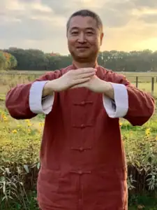 Man in a red traditional martial arts top stands in a sunlit field with yellow flowers, hands clasped at chest in a calm crossing pose