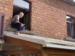 Older man in a striped shirt sits on a brick building's open window ledge, looking down at a small object in his hands.