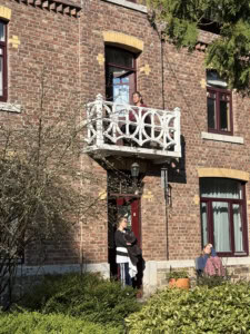 Two people stand by the front door of a red-brick building, with a white decorative balcony above them.