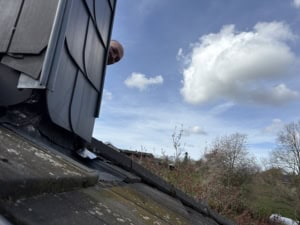 Person peeking over a dark slate roof from a dormer window, cloudy blue sky in the background.