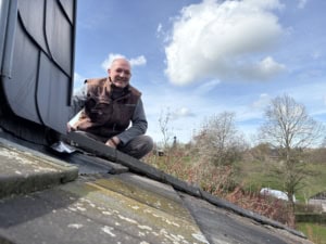 Man in a brown vest crouching on a roof edge, smiling at the camera with a blue sky and clouds behind him.