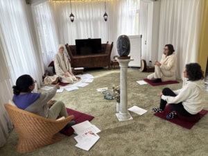 Participants sit cross-legged on red mats in a circle around a large blue stone sculpture on a pedestal; papers are scattered on the carpet.