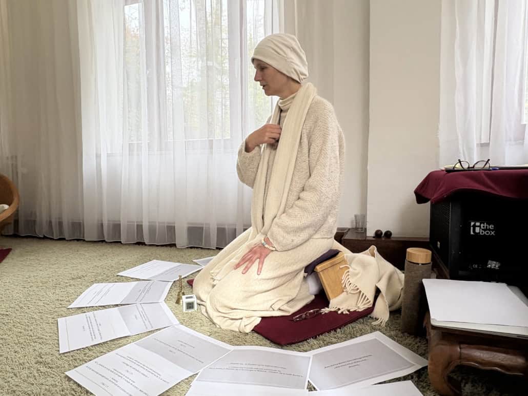 Person in a white robe and head wrap kneeling on a rug, surrounded by scattered papers in a bright room with sheer curtains.