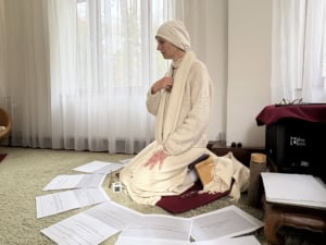 Person in a white robe and head wrap kneeling on a rug, surrounded by scattered papers in a bright room with sheer curtains.