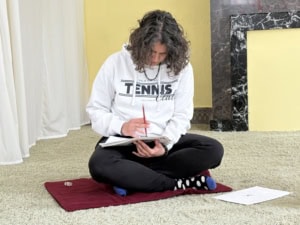 Person with curly hair in a white hoodie sits cross-legged on a red mat, writing in a notebook.