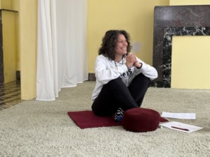 Woman with curly hair sits on a red cushion on a carpet, smiling and clasping hands, indoors near a yellow wall.