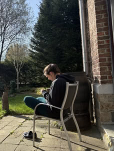 Person sitting on a patio chair outdoors, wearing a dark hoodie, with shoes off on the ground and greenery in the background.