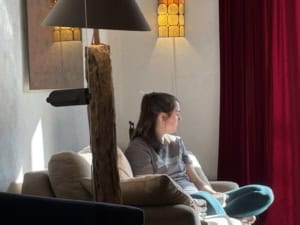Woman sitting on a beige sofa in a living room, with a tall wooden floor lamp and warm amber lamps, red curtains to the right.