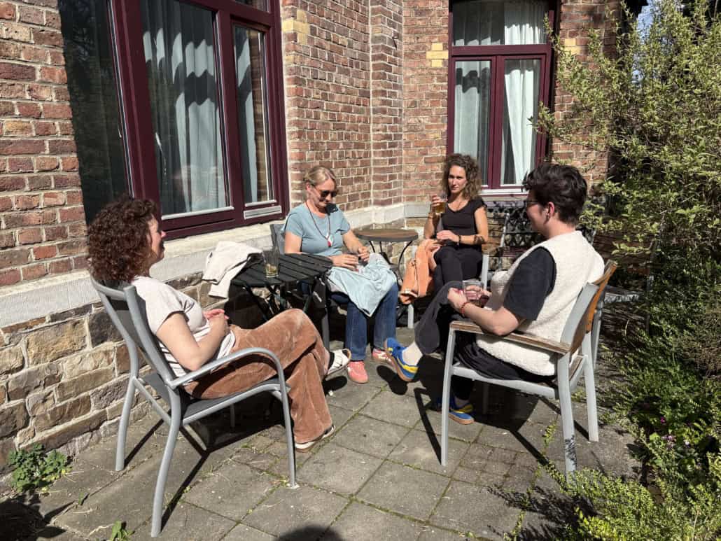 Four adults sit around a small table on a sunny brick patio, chatting with drinks in hand.