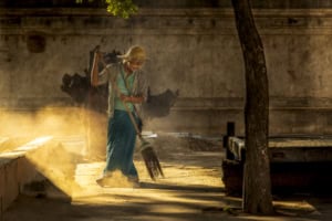 Person wearing a straw hat sweeps with a long broom in a smoky, sunlit courtyard beside a concrete wall.
