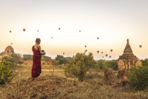 Monk in maroon robes holds a bowl while looking at hundreds of hot air balloons over ancient temple ruins at sunrise.