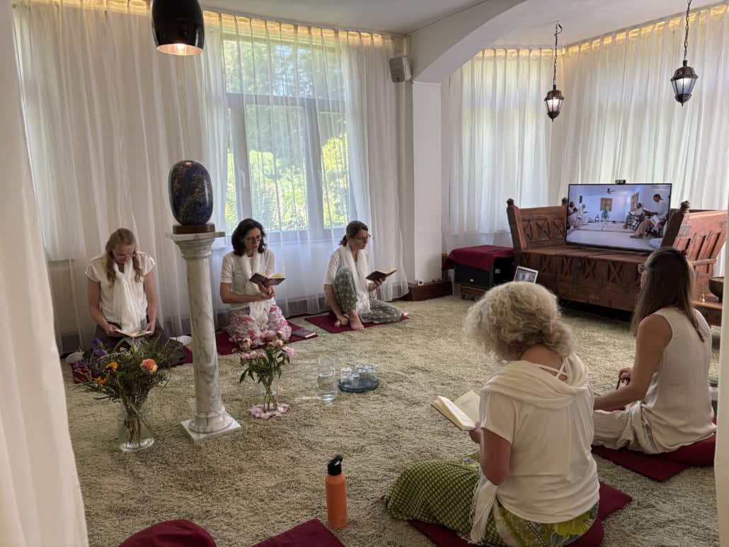 Women in white robes sit cross‑legged on cushions reading from small books in a bright, curtain‑lit room with flowers and a screen showing a video call or presentation.