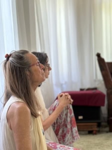 Side-profile of a woman wearing glasses, light sleeveless top, and a hair clip, hands clasped, seated indoors with white curtains in the background.