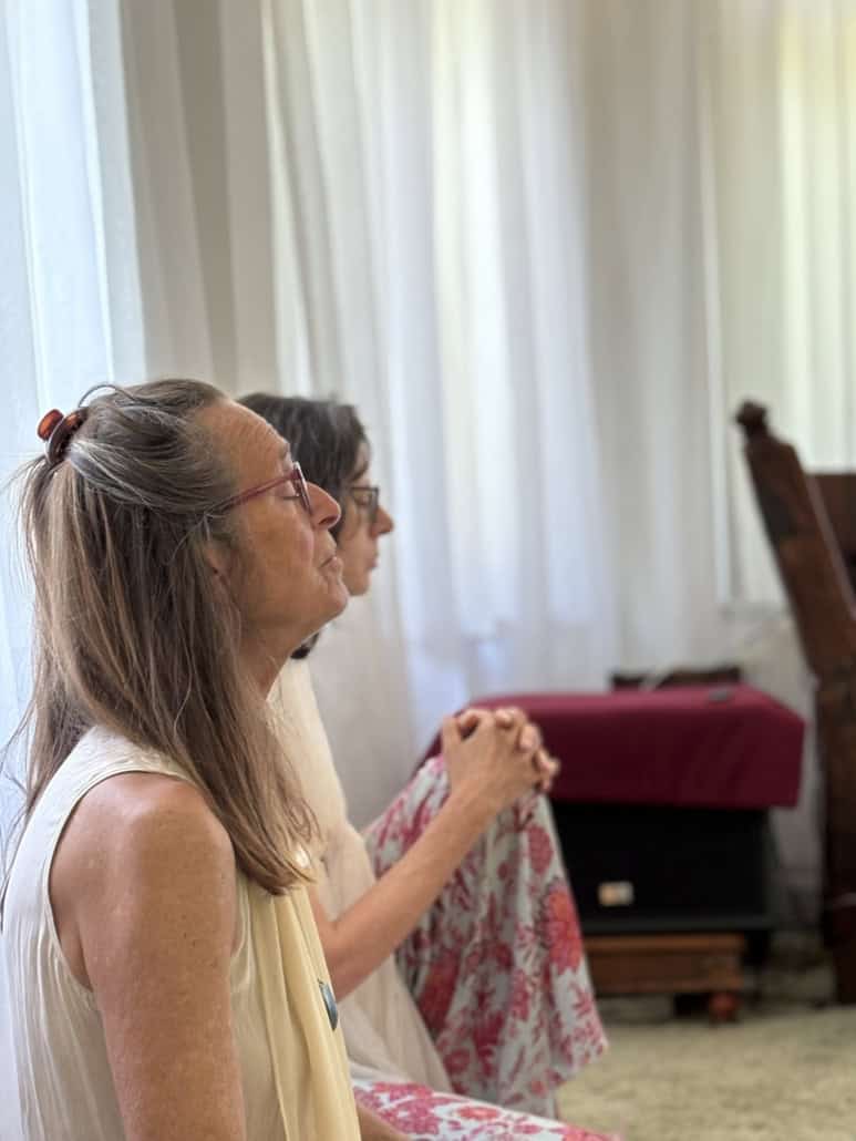 Side-profile of a woman wearing glasses, light sleeveless top, and a hair clip, hands clasped, seated indoors with white curtains in the background.