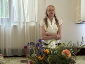 Woman in a white blouse with glasses stands with arms crossed in a bright room, flowers in the foreground.