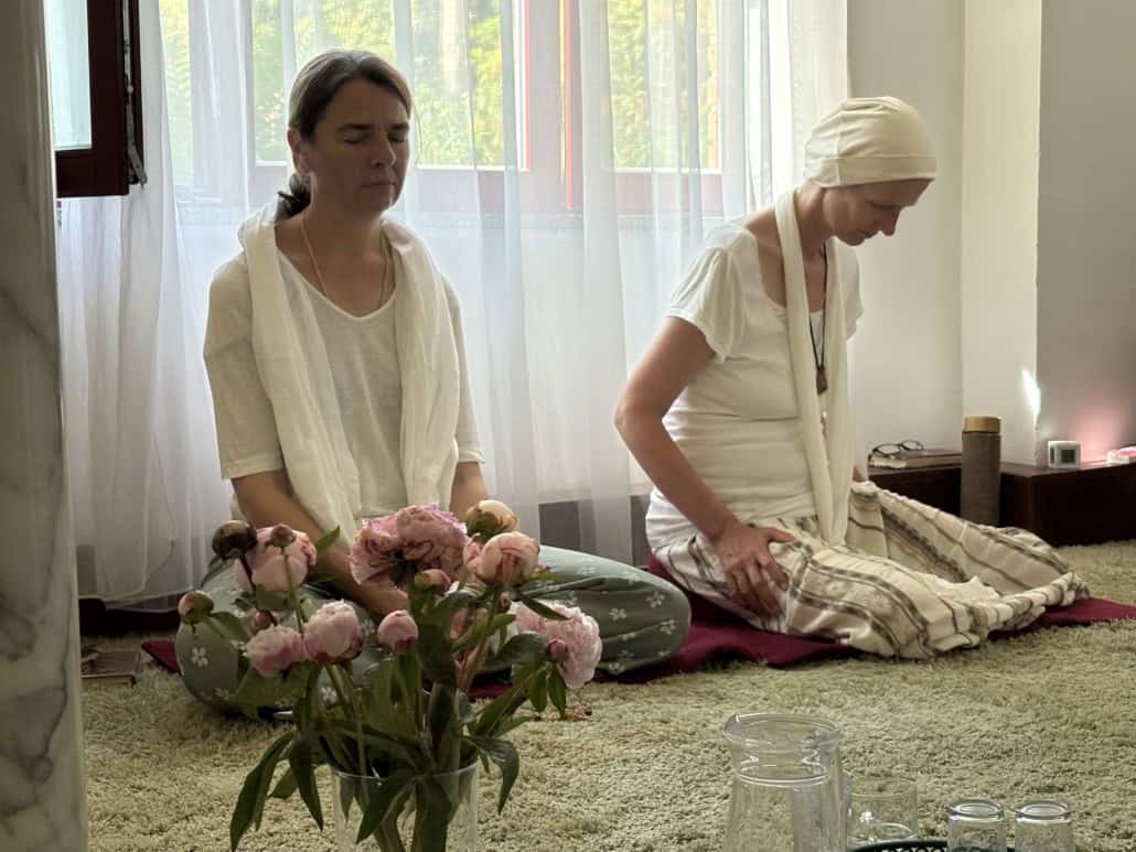 Two women in white clothing sit quietly on a carpeted floor, with a vase of pink peonies in the foreground and sheer curtains behind them.