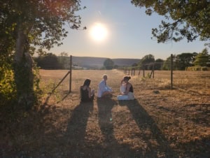 IMG_20260812_195400_612 | Asharum Amonines Group of people sitting on a dry field near a wooden fence as the sun shines brightly in a rural landscape, chatting and relaxed.