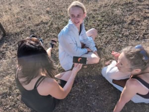 IMG_20260812_195440_535 | Asharum Amonines Group of friends sitting on dry grass outdoors, looking at a smartphone together with an orange crescent logo displayed on screen.