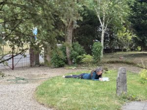 IMG_6042 2 | Asharum Amonines Person lying on the grass outdoors, writing in a notebook near a garden and trees.