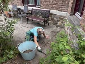 IMG_6107 2 | Asharum Amonines Person kneeling on a stone patio, gardening with a blue bucket nearby, by a brick wall and bench setup.