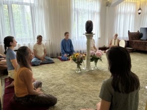 IMG_6309 | Asharum Amonines Group of people seated cross-legged on cushions in a sunlit room, meditating around a marble pedestal with a dark stone sculpture, flowers in front.