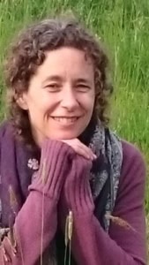 Woman with curly hair smiling outdoors in a grassy field, wearing a maroon sweater and scarf.