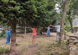 Group of people standing in a circle outdoors under a large evergreen tree, arms extended as they practice a group exercise.