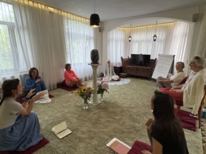 Group of people seated on red cushions in a bright room, forming a circle around a flower arrangement.